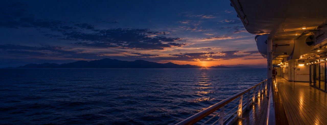 A ship’s deck at dusk, the railing leading toward a glowing sunset over the sea and distant hills